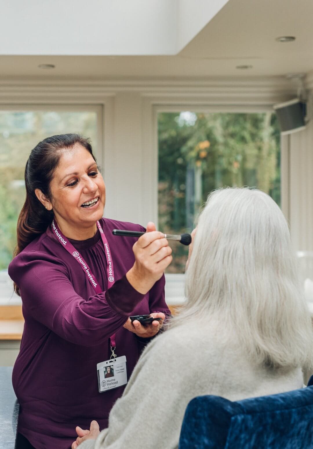 A caregiver applies makeup to an elderly woman seated in a well-lit room with large windows. - Home Instead