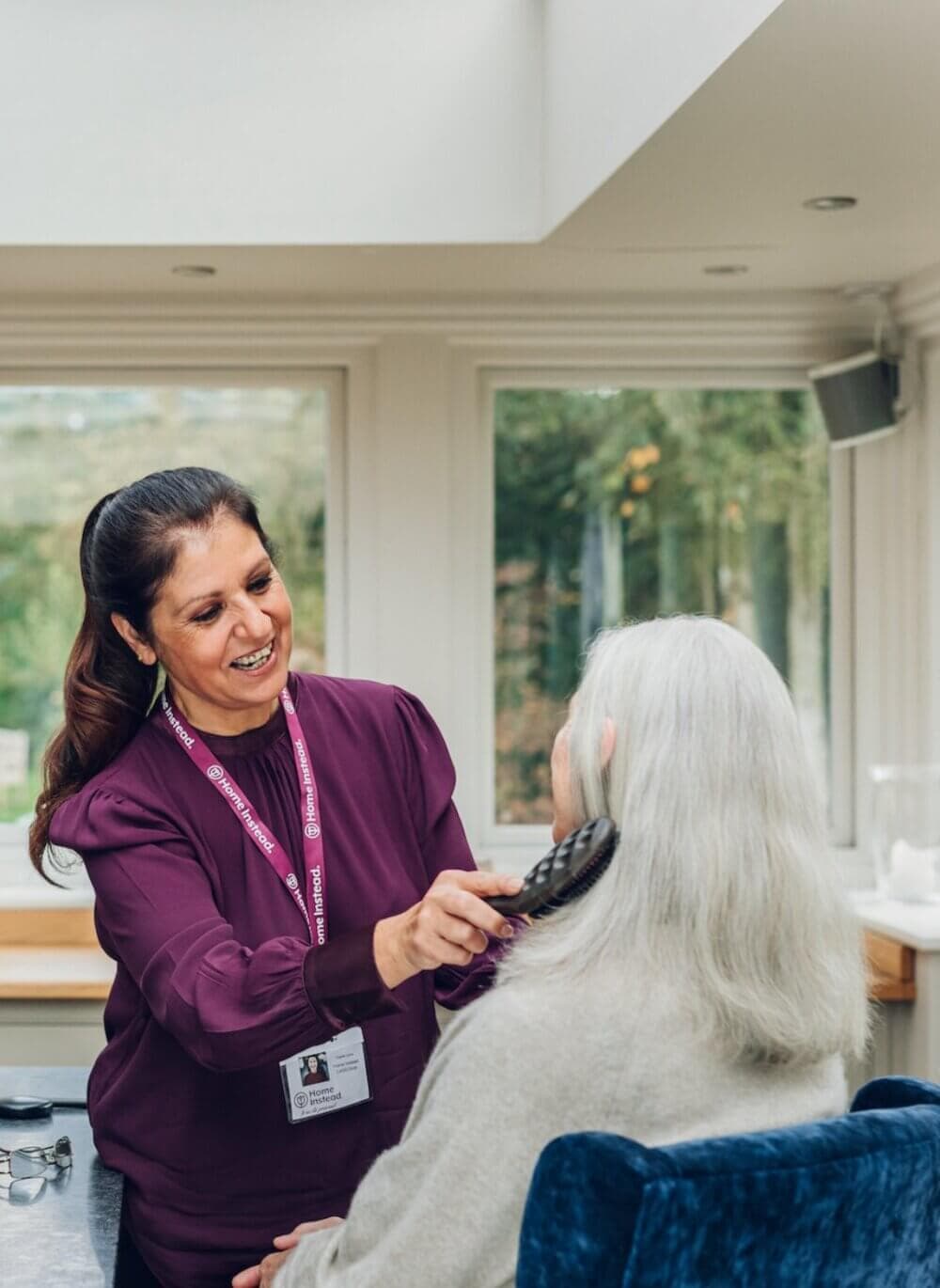 A woman in a purple shirt brushes the hair of an older woman with gray hair in a bright room with large windows. - Home Instead