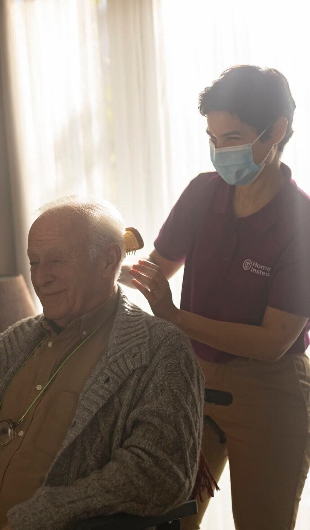 A caregiver wearing a mask combs the hair of an elderly man in a wheelchair, both smiling warmly. - Home Instead