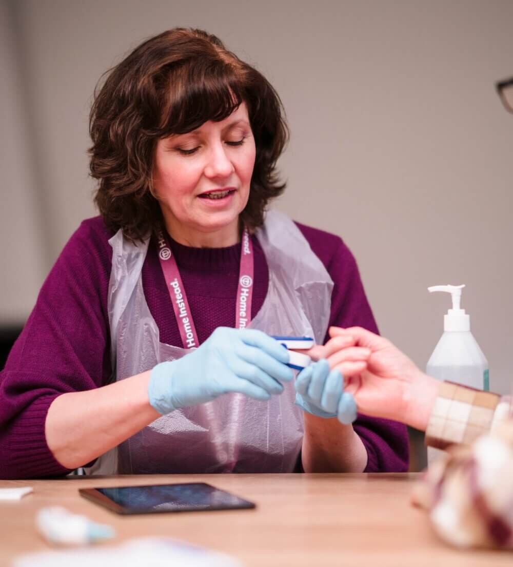 Healthcare worker wearing gloves and apron performing a finger prick test on a person’s hand at a table. - Home Instead