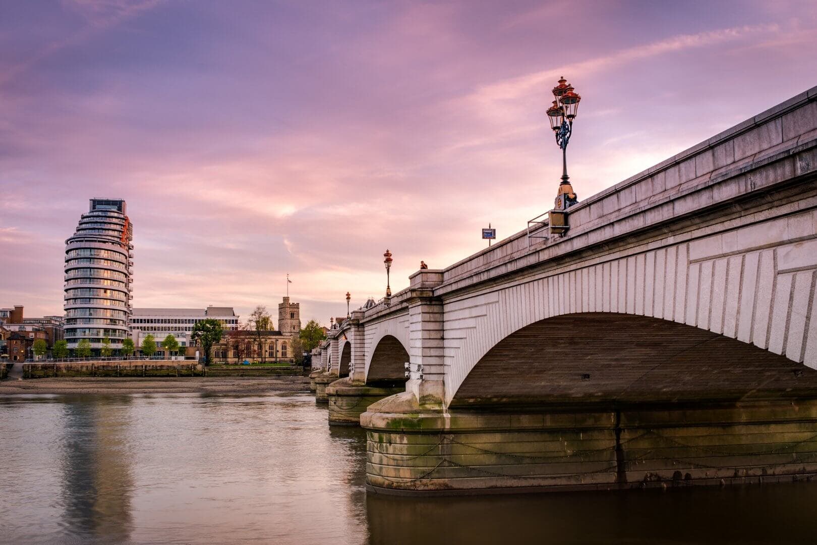 Bridge over calm river with modern and historic buildings in the background at sunset. Stunning sky with pink hues. - Home Instead