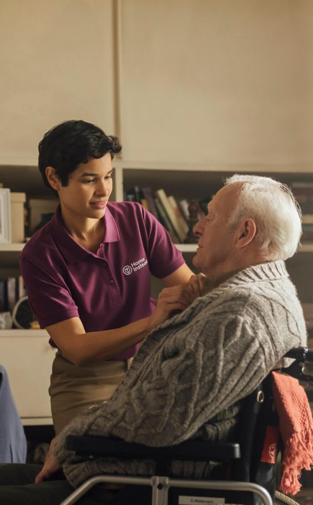 A caregiver in a burgundy shirt helps an elderly man in a wheelchair with his sweater inside a home. - Home Instead