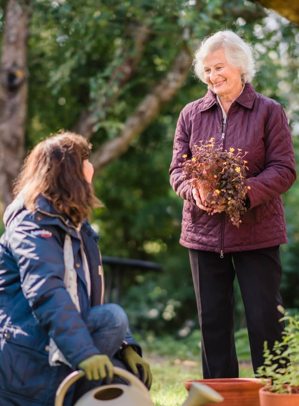 Elderly woman holding a plant, smiles at a younger woman kneeling with gardening tools in a garden. - Home Instead