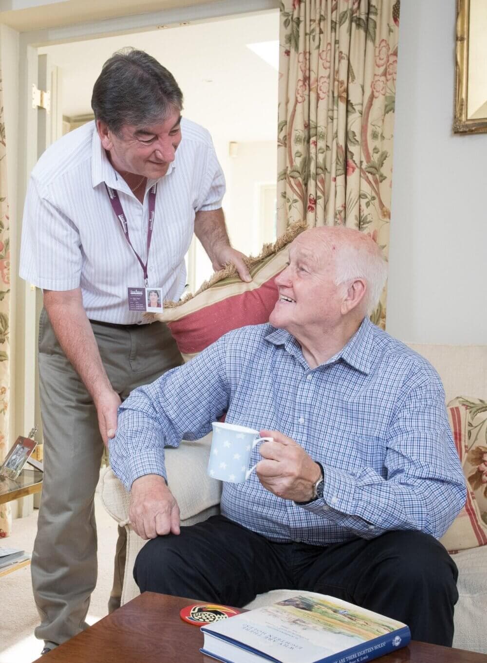 A caregiver interacts warmly with an elderly man holding a mug, seated in a cozy, well-lit living room. - Home Instead