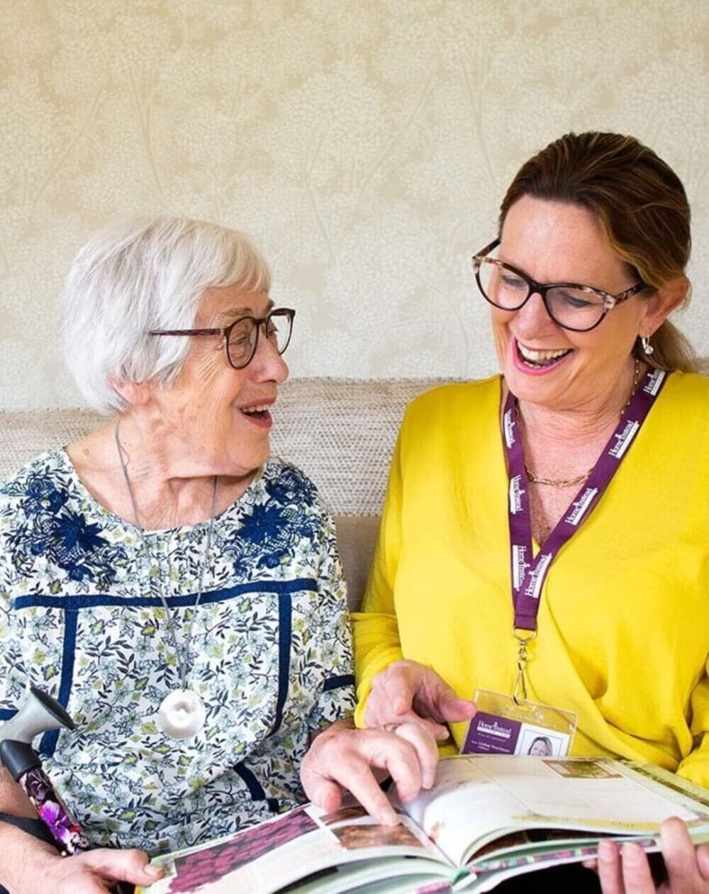 An elderly woman and a young woman happily look at a photo album while sitting on a sofa in a well-lit room. - Home Instead