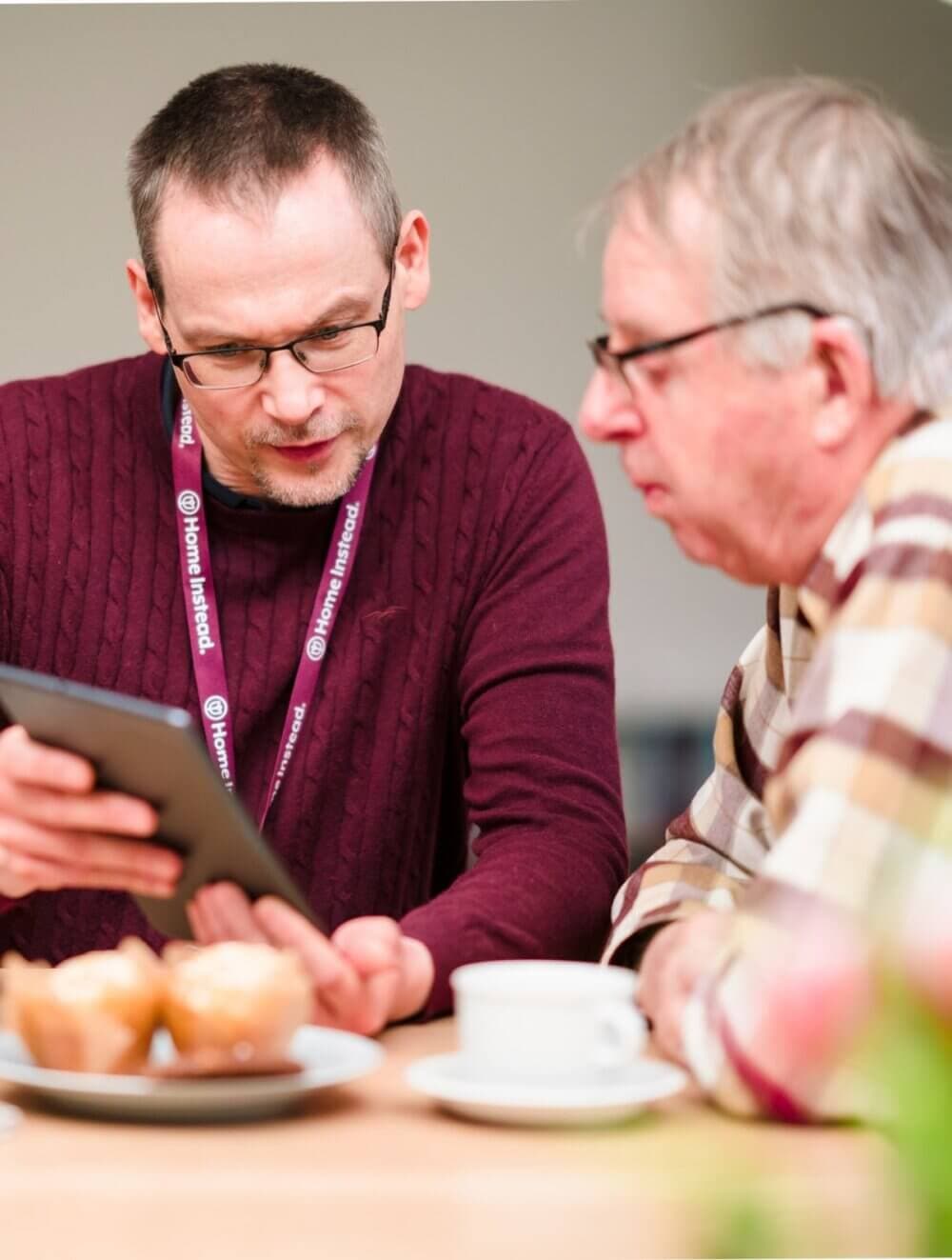 Two men sitting at a table, with one man holding a tablet and showing it to the other. Coffee cups and pastries are on the table. - Home Instead Bournemouth & Christchurch