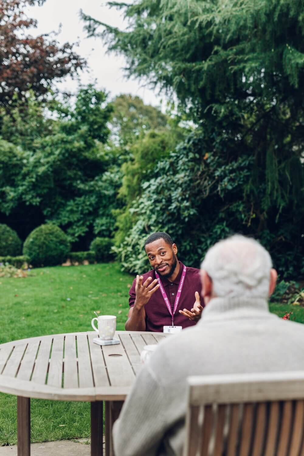 A Care Professional in the garden talking to an older person at an outdoor table with trees and bushes in the background. - Home Instead Bournemouth & Christchurch