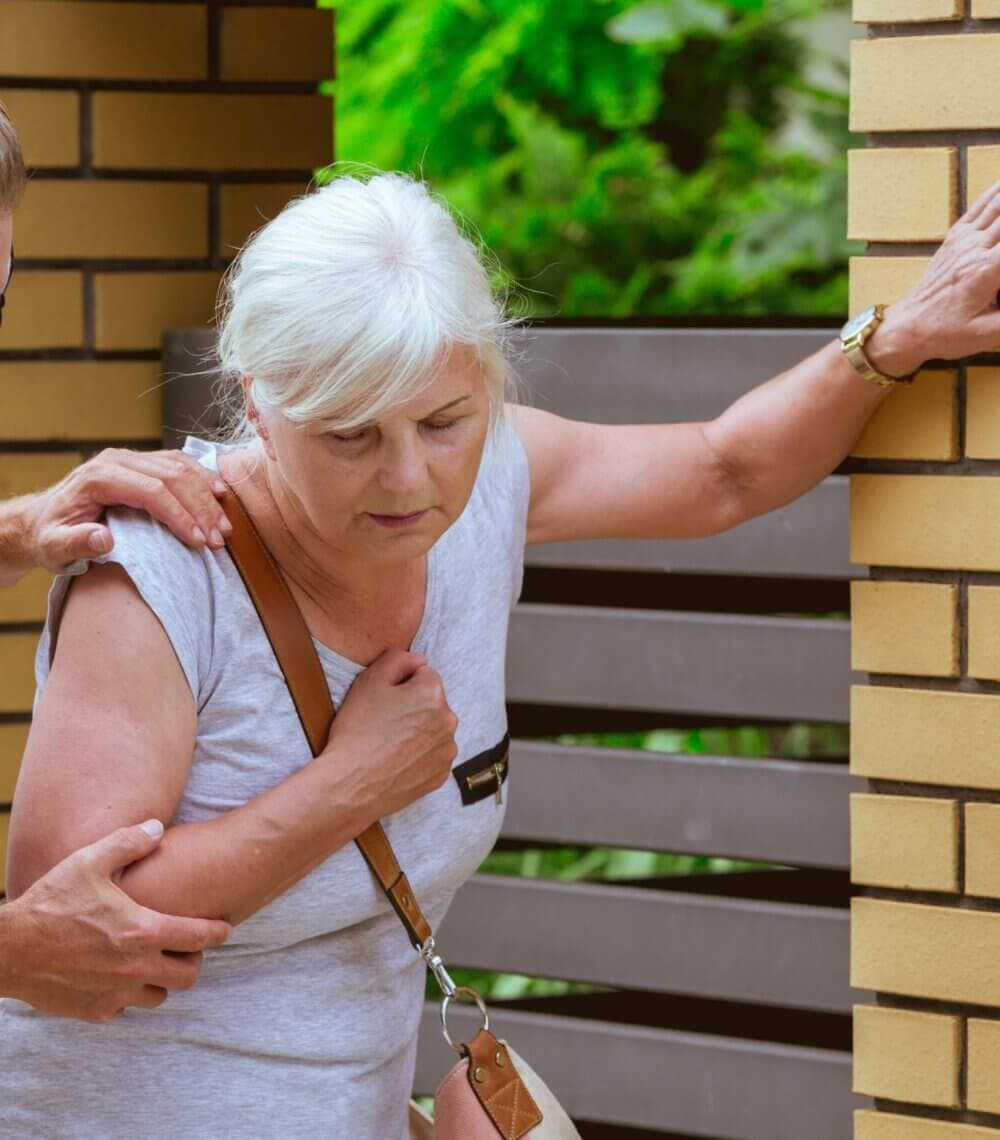 An older woman looking unwell is being supported by someone as she holds onto a brick wall for balance. - Home Instead