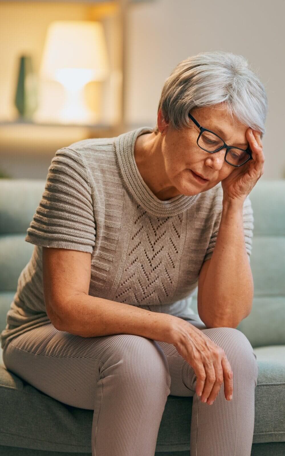 An elderly woman with short grey hair and glasses sits on a couch, holding her head and looking distressed. - Home Instead Bournemouth & Christchurch
