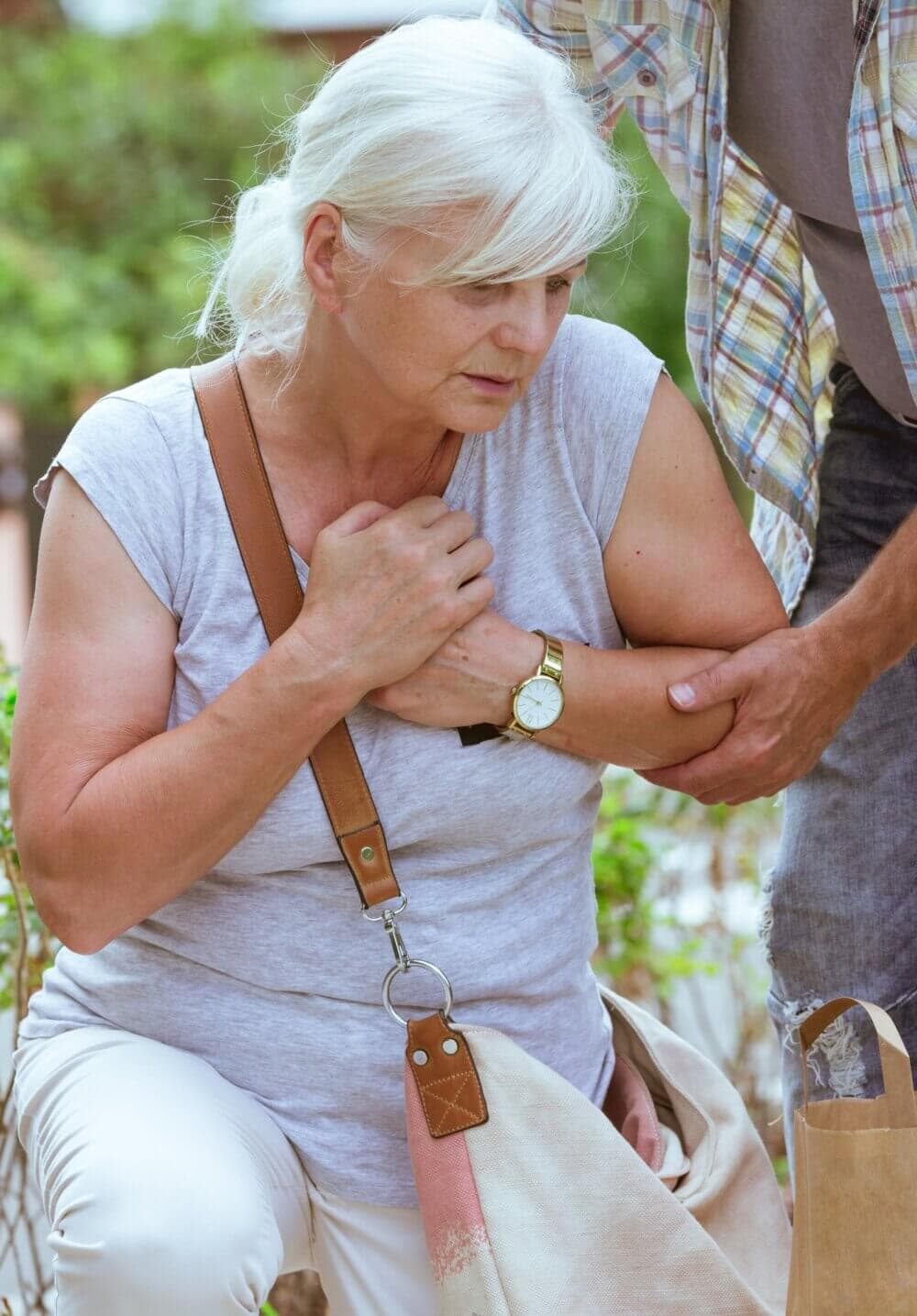 An elderly woman clutching her chest is assisted by a man. She is wearing a light gray shirt and has a shoulder bag. - Home Instead