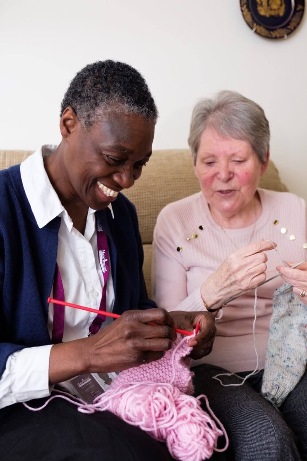 Two women sitting on a couch, knitting together and smiling. - Home Instead