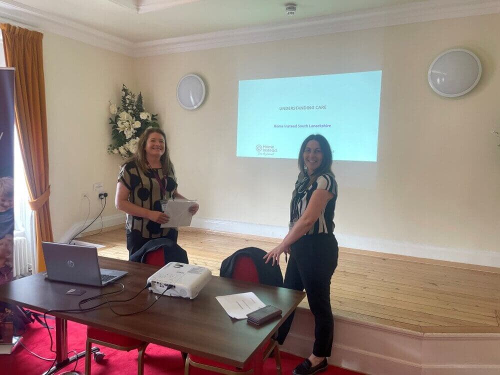 Two people smiling in a classroom, standing near a table with a laptop and projector displaying "Understanding Care. - Home Instead