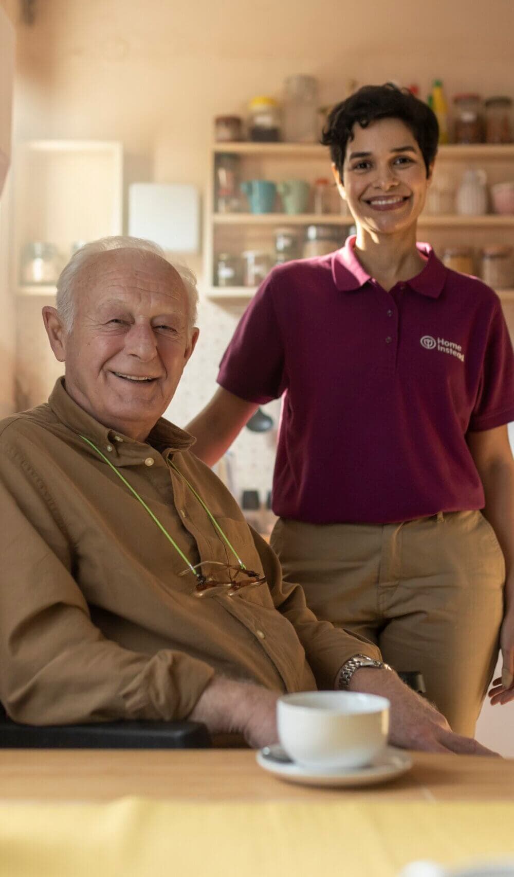 Senior man sitting at a table with a caregiver standing beside him, both smiling, indoors with kitchen items in the background. - Home Instead