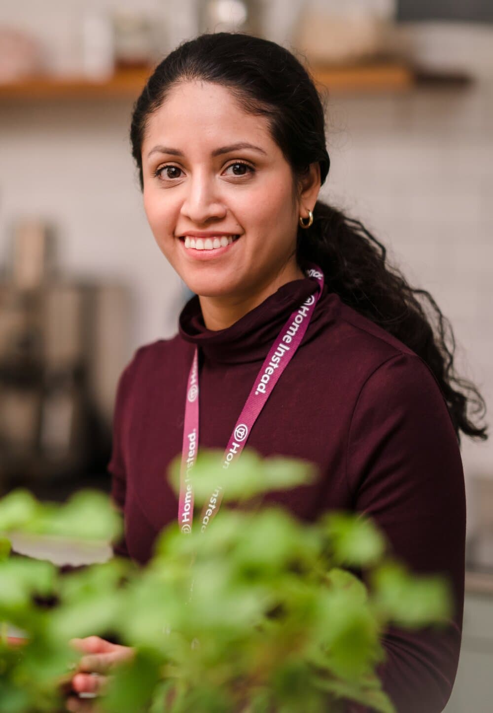 Woman with dark hair wearing a maroon top and lanyard, smiling indoors with plants in the foreground. - Home Instead