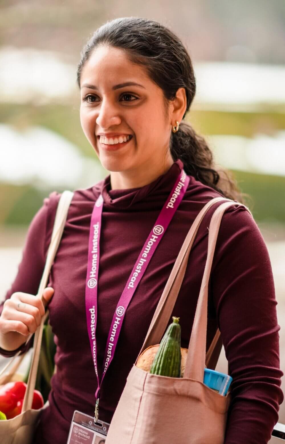 A smiling woman wearing a purple shirt and a lanyard sits at a table with a teapot and cups. - Home Instead
