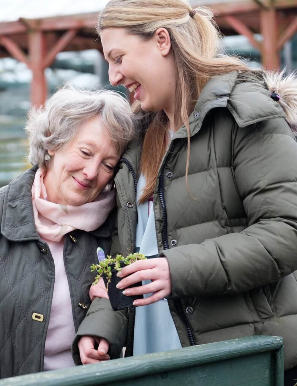 An older woman and a younger woman smile while holding a small plant. The older woman rests her head on the younger's shoulder. - Home Instead Poole