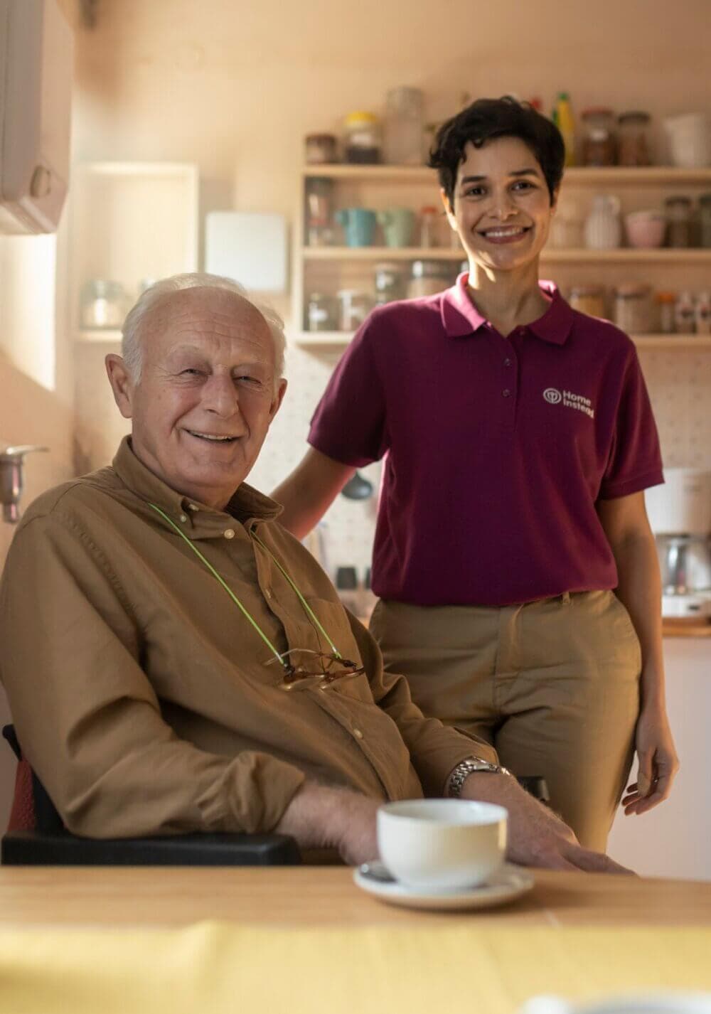 An elderly man sits at a table with a caregiver standing beside him, both smiling, in a cozy kitchen with shelves. - Home Instead