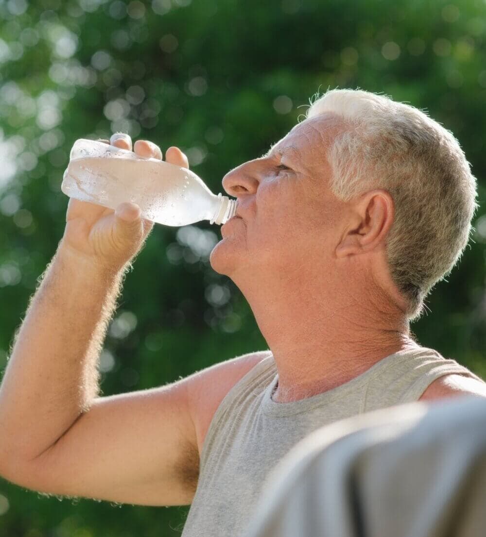 Older man drinking water from a bottle outdoors, with a blurred green background. - Home Instead Bournemouth & Christchurch