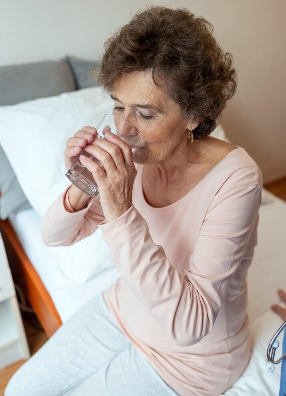 An elderly woman with curly hair sits on a bed and drinks a glass of water. - Home Instead
