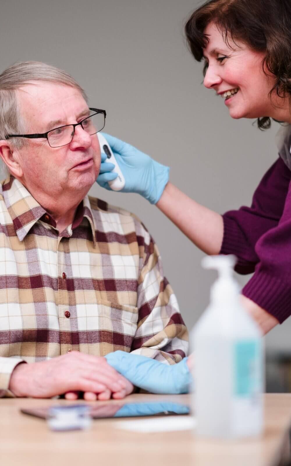 A woman wearing gloves helps an older man with glasses by holding a cloth or device to his face. - Home Instead