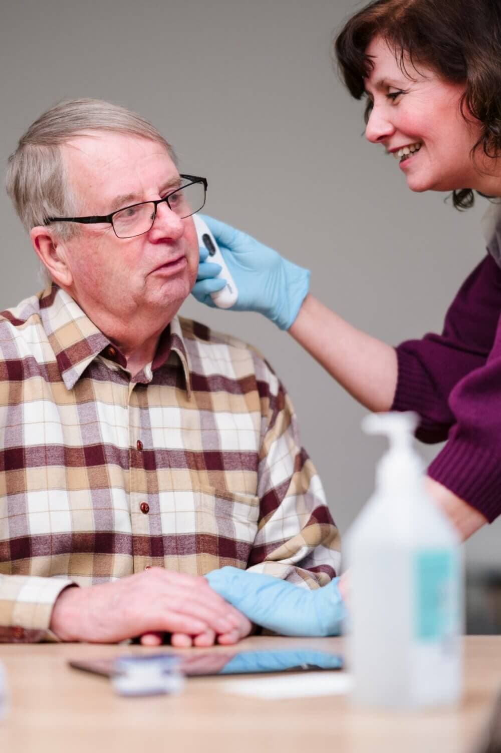 A caregiver in gloves checks an elderly man's ear with an otoscope. They both appear to be smiling. - Home Instead