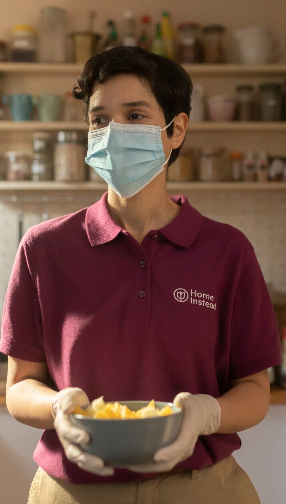 Person in maroon "Home Instead" shirt, face mask, and gloves holding a bowl in a kitchen setting. - Home Instead