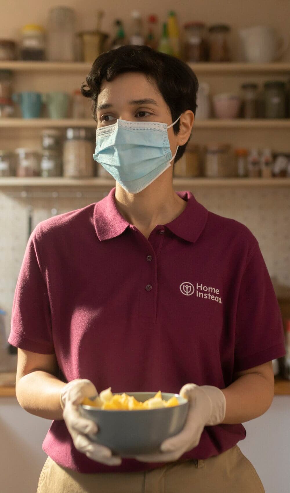 A person wearing a mask and gloves holds a bowl of food in a kitchen, wearing a "Home Instead" polo shirt. - Home Instead
