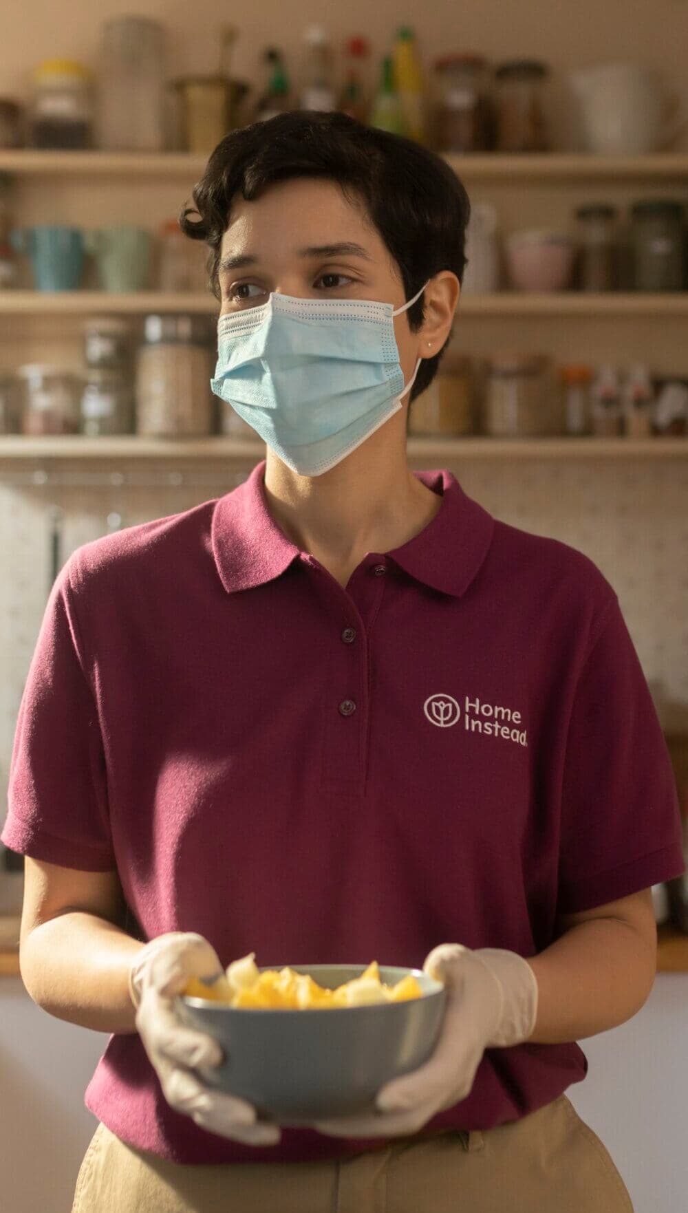 A person wearing a mask and gloves, in a maroon "Home Instead" uniform, holding a bowl in a kitchen. - Home Instead