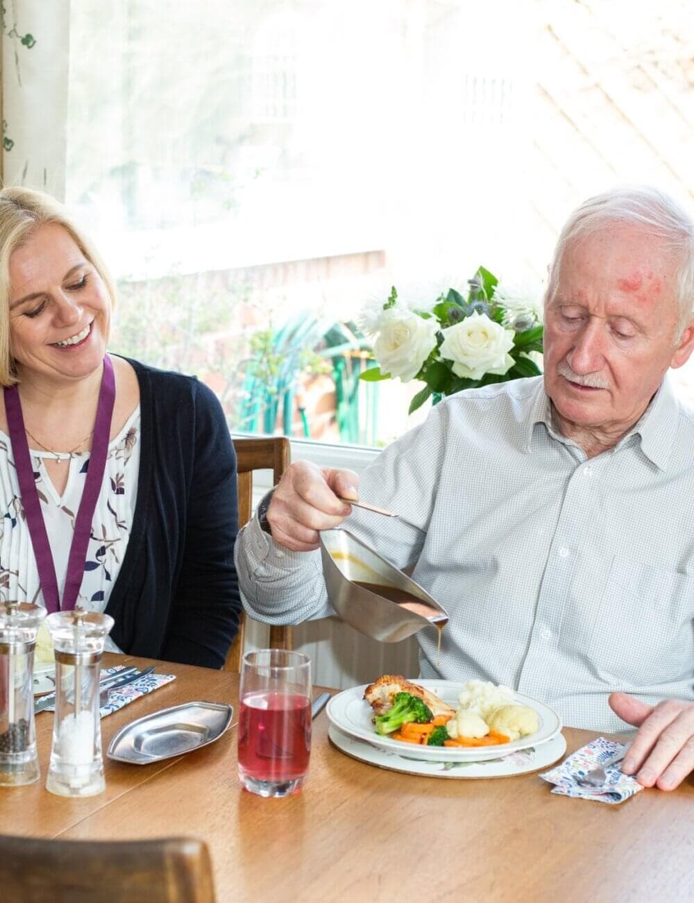 A smiling woman sits next to an older man pouring gravy onto his meal at a dining table. - Home Instead
