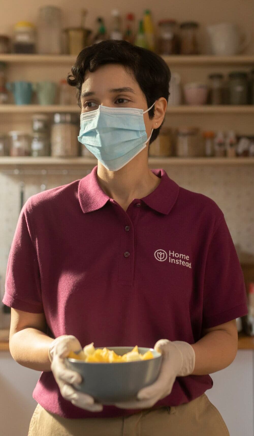 Person in a Home Instead shirt holding a bowl of food, wearing a surgical mask and gloves in a kitchen setting. - Home Instead