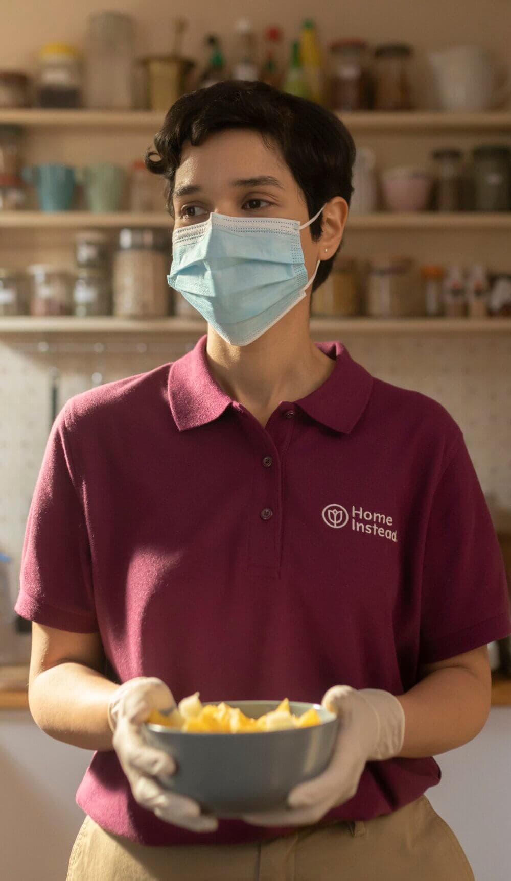 Person wearing a mask, gloves, and a maroon polo holding a bowl. Shelves with various jars in the background. - Home Instead