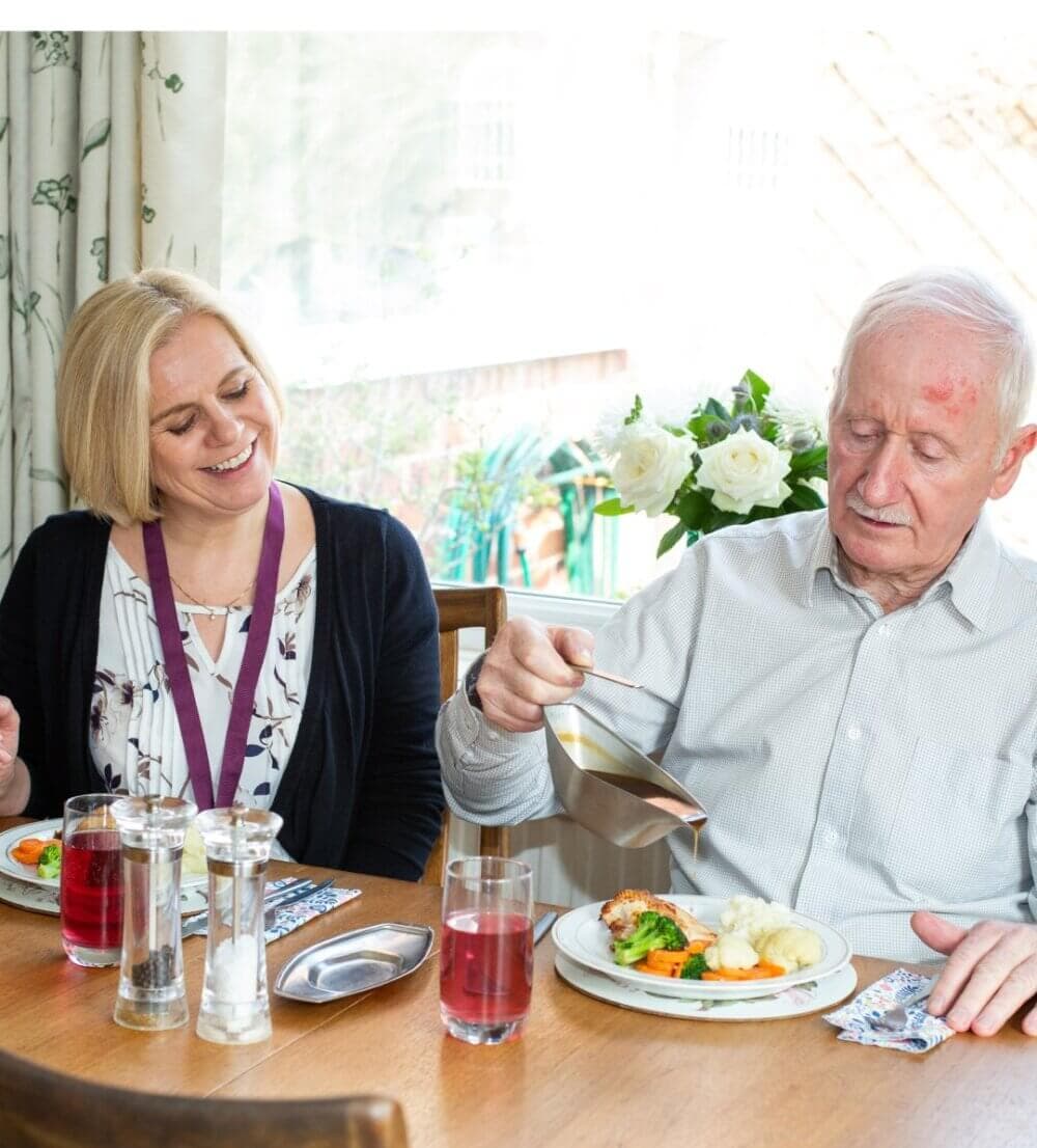 A smiling woman with a badge watches an older man pour sauce over his meal at a dining table. - Home Instead