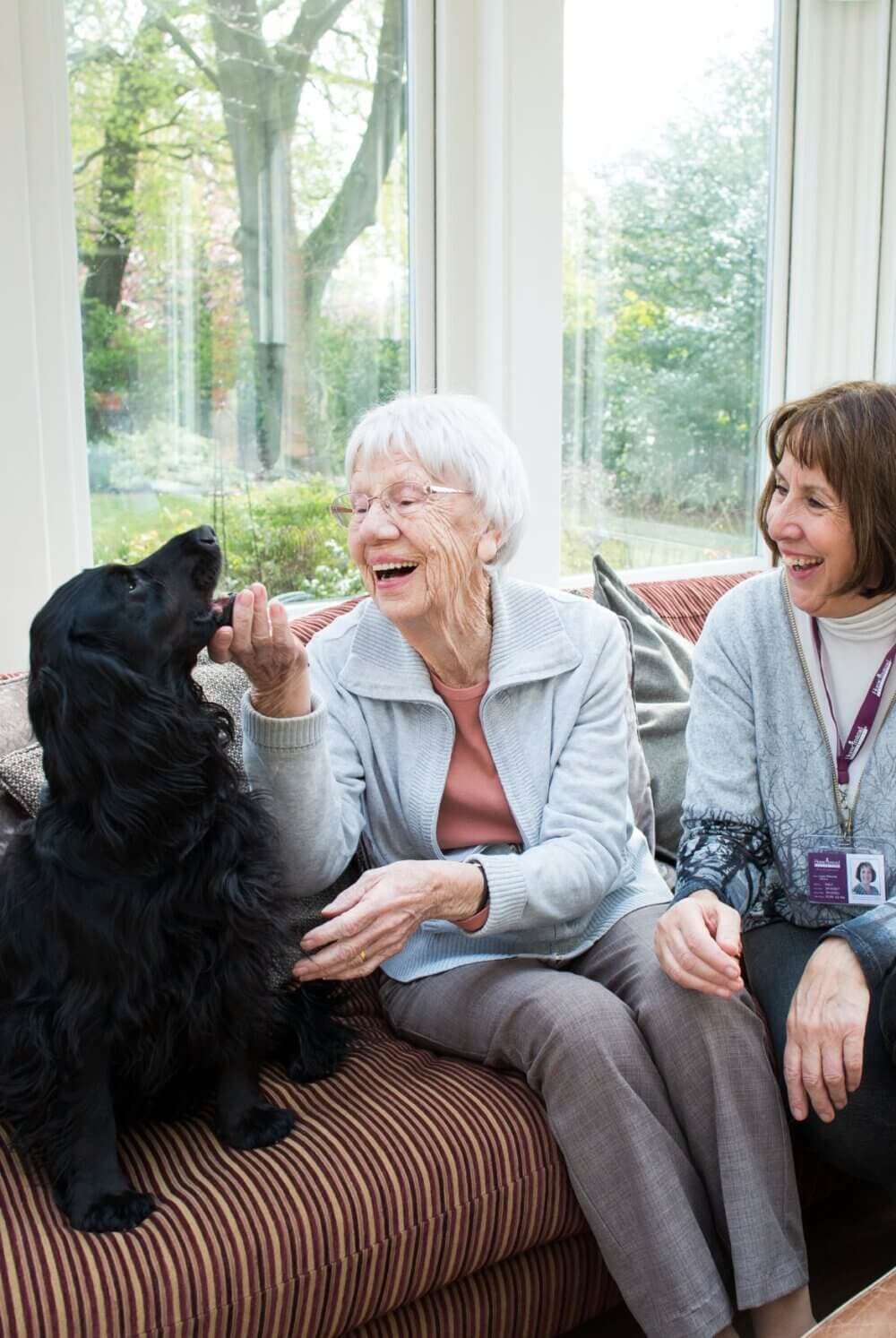 An elderly woman joyfully pets a black dog while sitting on a couch, accompanied by a smiling caregiver. - Home Instead