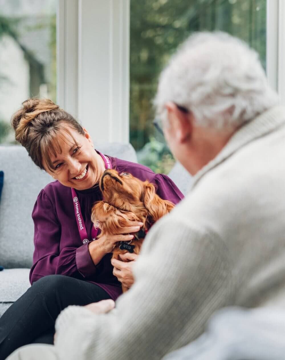 A smiling woman holds a dog while talking to an older man in a cozy, well-lit room. - Home Instead