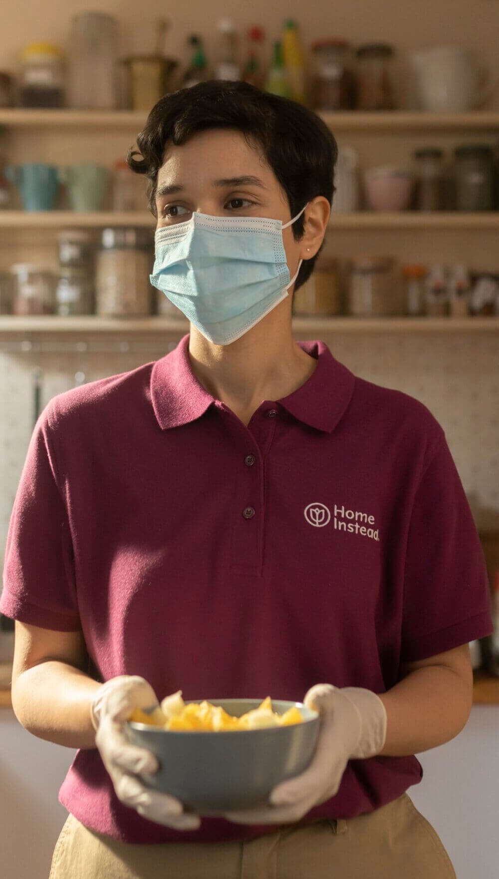 Person wearing a mask and gloves, holding a bowl, in a kitchen. Dressed in a "Home Instead" maroon shirt. - Home Instead