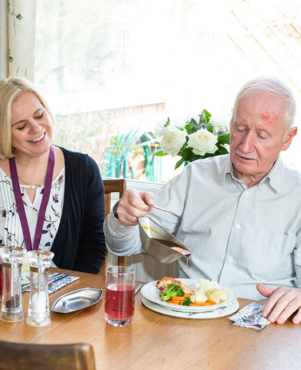 An elderly man is pouring gravy over his meal, while a smiling woman sits next to him at a dining table. - Home Instead