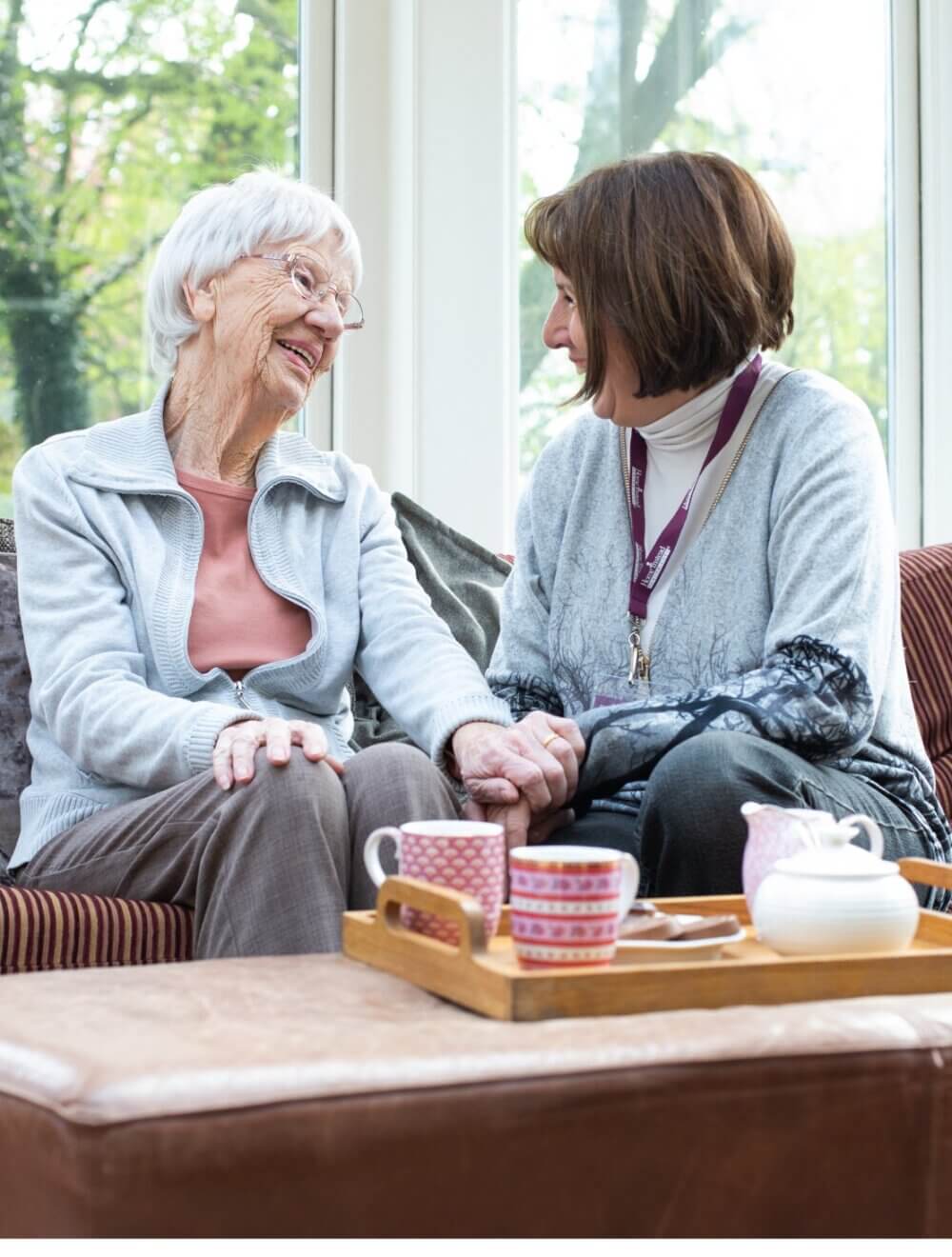 An elderly woman and a younger woman share a warm moment on a sofa, with tea on a tray in front of them. - Home Instead