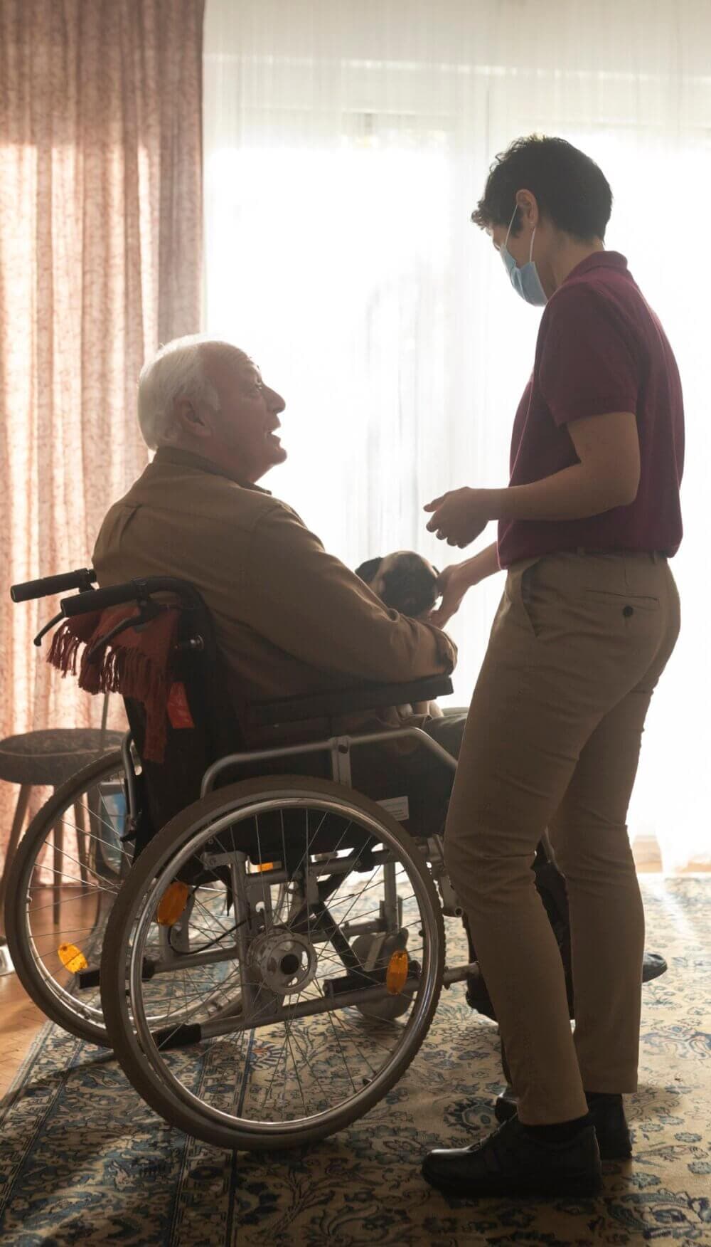 An elderly man in a wheelchair holds a small dog while a caregiver in a mask interacts with them in a warmly lit room. - Home Instead