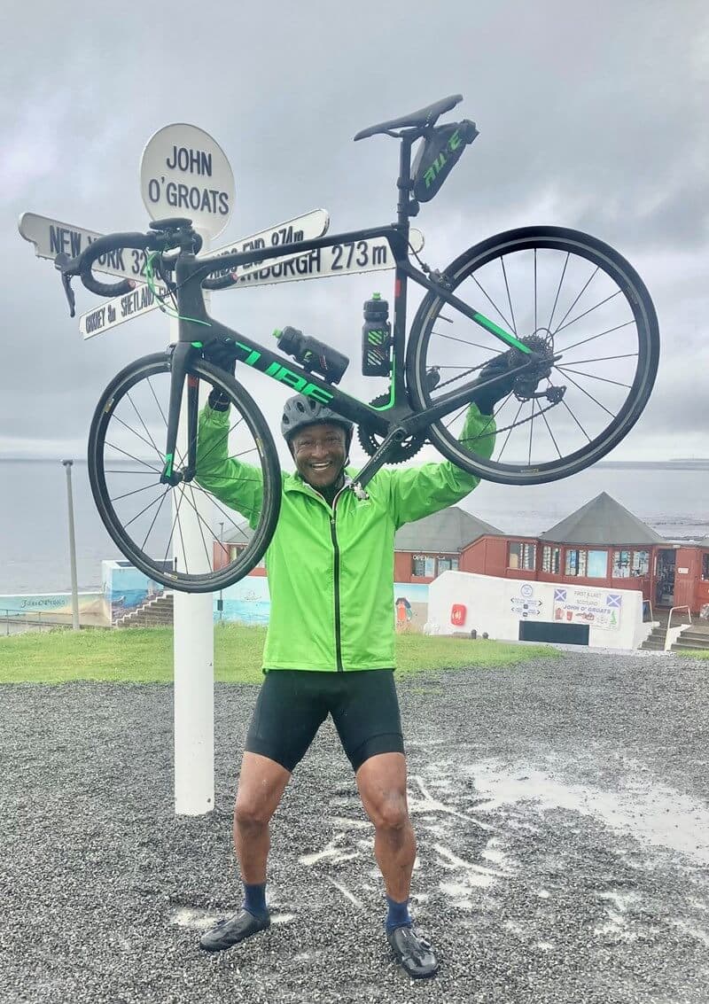 Cyclist in a green jacket lifts bike overhead at John O'Groats signpost on a cloudy day in a coastal area. - Home Instead