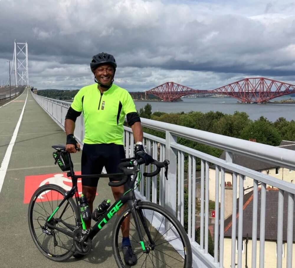 A cyclist in a neon green jersey stands with a bicycle on a bridge with red rail bridges in the background. - Home Instead