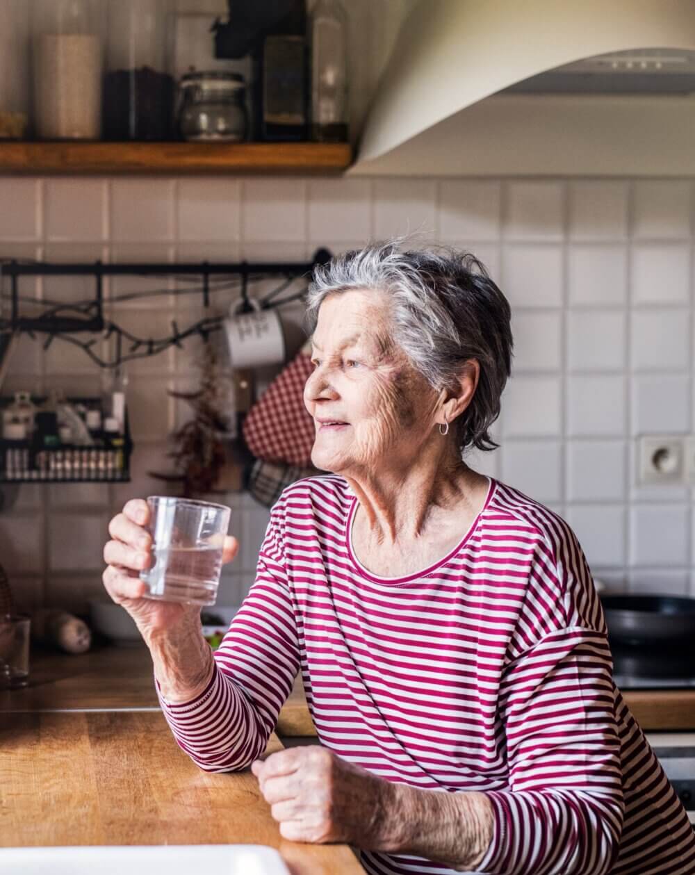 Elderly woman in a red and white striped shirt holding a glass of water, sitting in a kitchen, smiling and looking away. - Home Instead