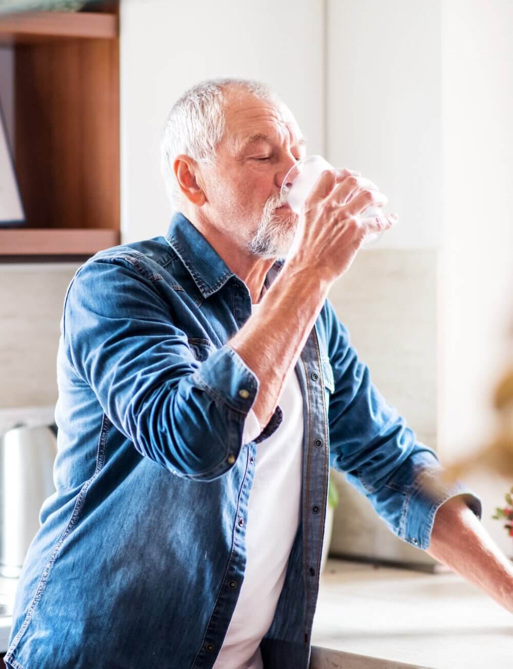 Elderly man in a denim shirt drinking a glass of water in a bright kitchen. - Home Instead Bournemouth & Christchurch