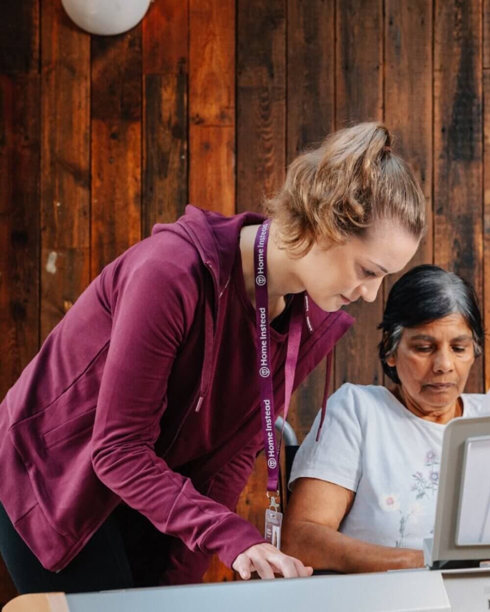 A woman in a purple jacket helps another woman with a laptop, against a wooden wall background. - Home Instead