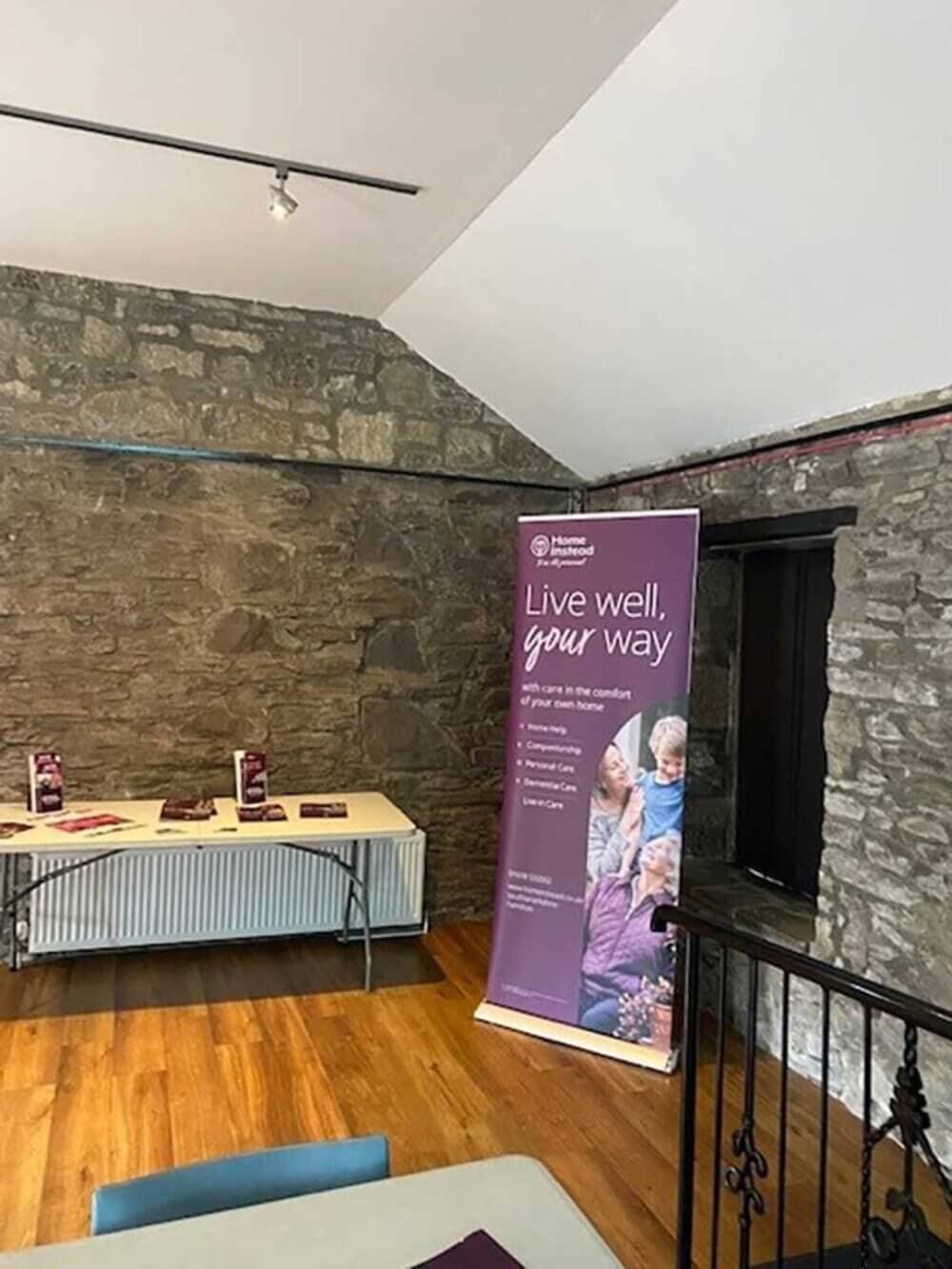 A room with a stone wall, wooden floor, table with brochures, and a standing banner that reads "Live well, your way. - Home Instead