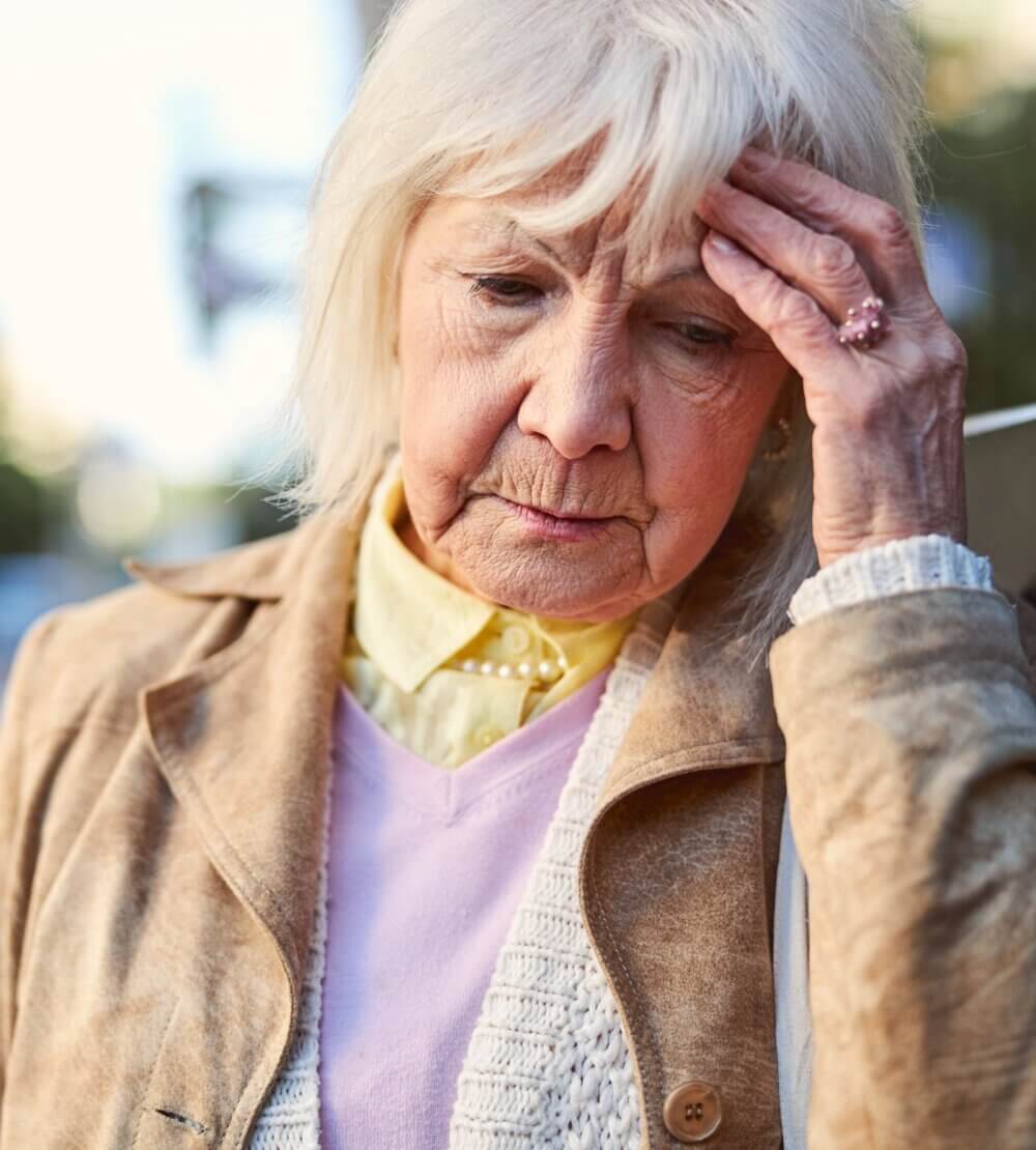 Elderly woman with gray hair looks worried while resting her hand on her forehead, wearing a beige coat and light sweater. - Home Instead
