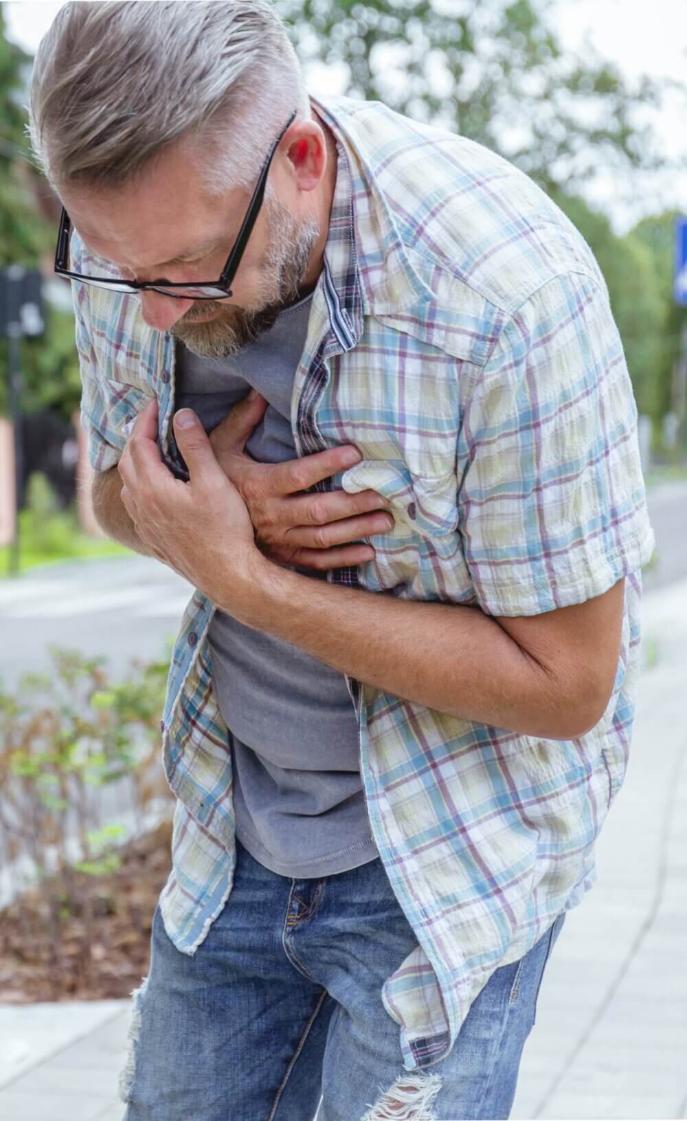 A man with glasses and a gray beard clutching his chest while walking outdoors, appearing to be in pain. - Home Instead