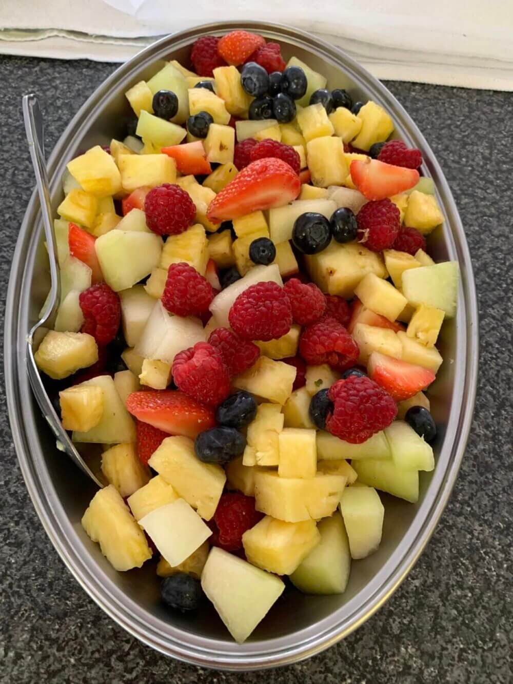 A metal bowl filled with a colorful fruit salad containing pineapple, strawberries, raspberries, blueberries, and melon. - Home Instead