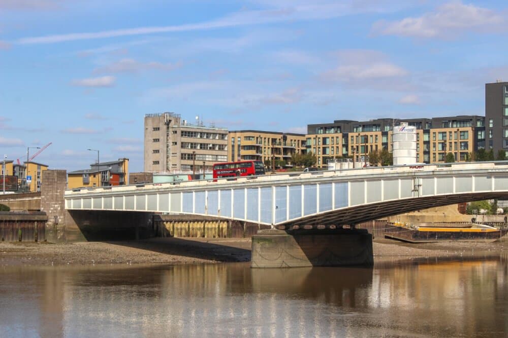 A red double-decker bus crossing a bridge over a river, with modern buildings and a crane in the background on a clear day. - Home Instead