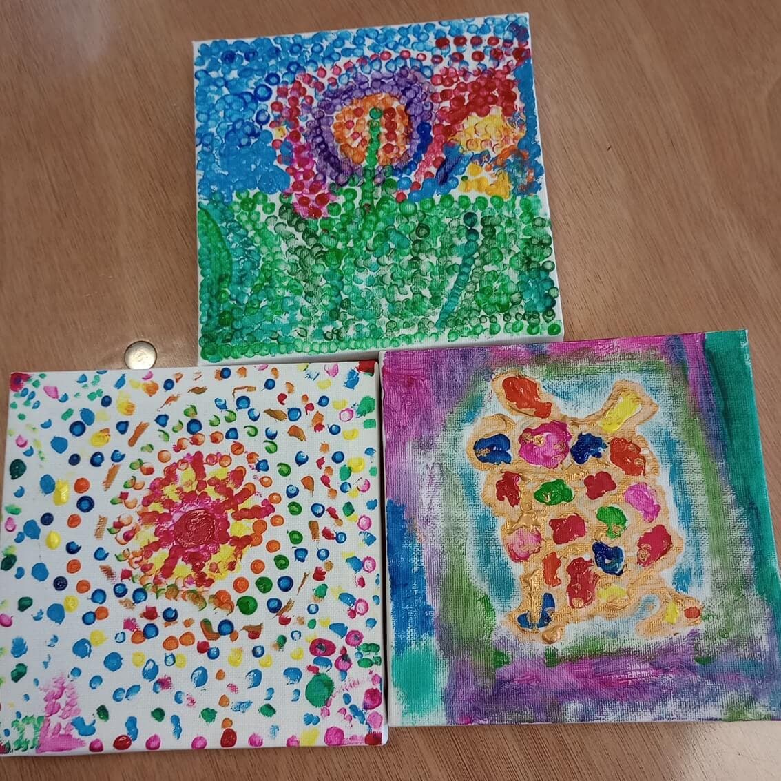 Three colorful, abstract paintings on a table featuring dot patterns and vibrant designs of a snail, a flower, and a bunny. - Home Instead