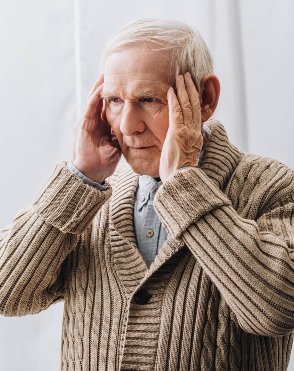 Elderly man in a knitted sweater holding his temples, looking concerned. - Home Instead